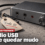 Ilustración con relieve sobre una losa de hormigón pulido gris cálido con los poros y el árido visibles bajo una luz rasante: en el centro, una interfaz de audio USB de escritorio con chasis de aluminio cepillado grafito, apoyada en tres cuartos, con el canto frontal de unos cuatro centímetros a la vista, una perilla de volumen moleteada girada al mínimo y un LED de señal redondo apagado; del aparato sale un cable USB trenzado que cae flojo sobre el hormigón, y de ese cable cuelga hacia el frente una etiqueta de papel rígido color hueso con ojal metálico donde dice a mano «Code 10», con su sombra sobre la piedra; detrás y a la izquierda, una chapita circular con el logo de cuatro cuadrados de Microsoft apoyada boca arriba; en la banda inferior, el antetítulo «Parche de septiembre» y el titular «El audio USB puede quedar mudo»