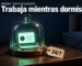 Ilustración: de noche, sobre una mesa de luz de nogal, una campana de vidrio encierra una computadora portátil en miniatura encendida, cuya pantalla verde es la única luz de la escena; atada a la base, una etiqueta de cartulina dice «24/7». Arriba, en una franja azul oscuro, el logo de OpenAI, el texto «OpenAI · dots en ChatGPT» y el titular «Trabaja mientras dormís».